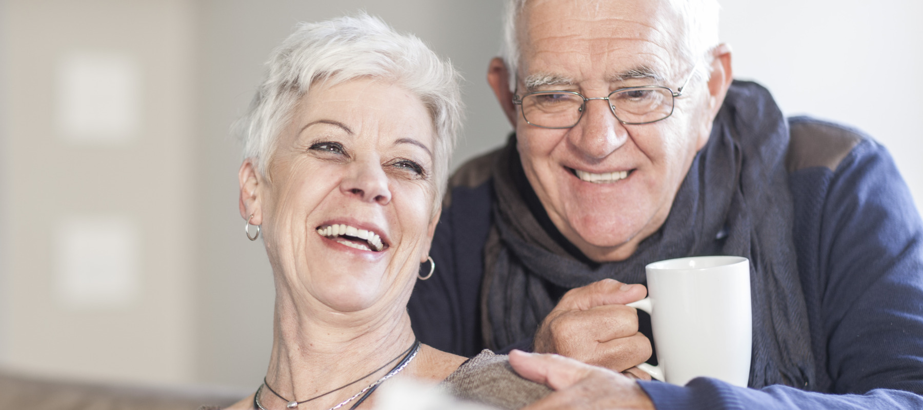 A happy senior couple enjoys a quiet day at home, with a man leaning over the back of the couch to look at a newspaper with his wife.