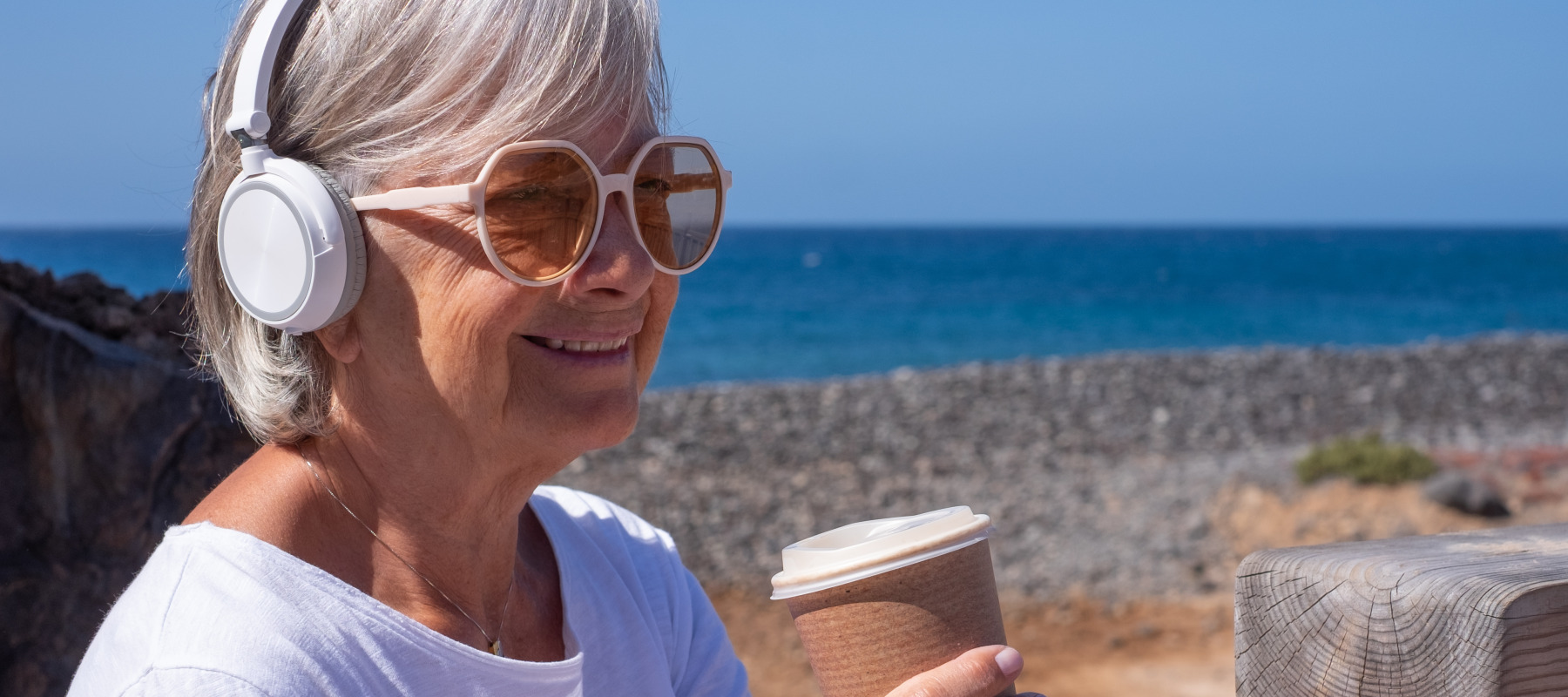 A senior woman with short, wavy grey hair wearing white headphones and large tinted glasses smiles as she holds a disposable coffee cup on a beach.