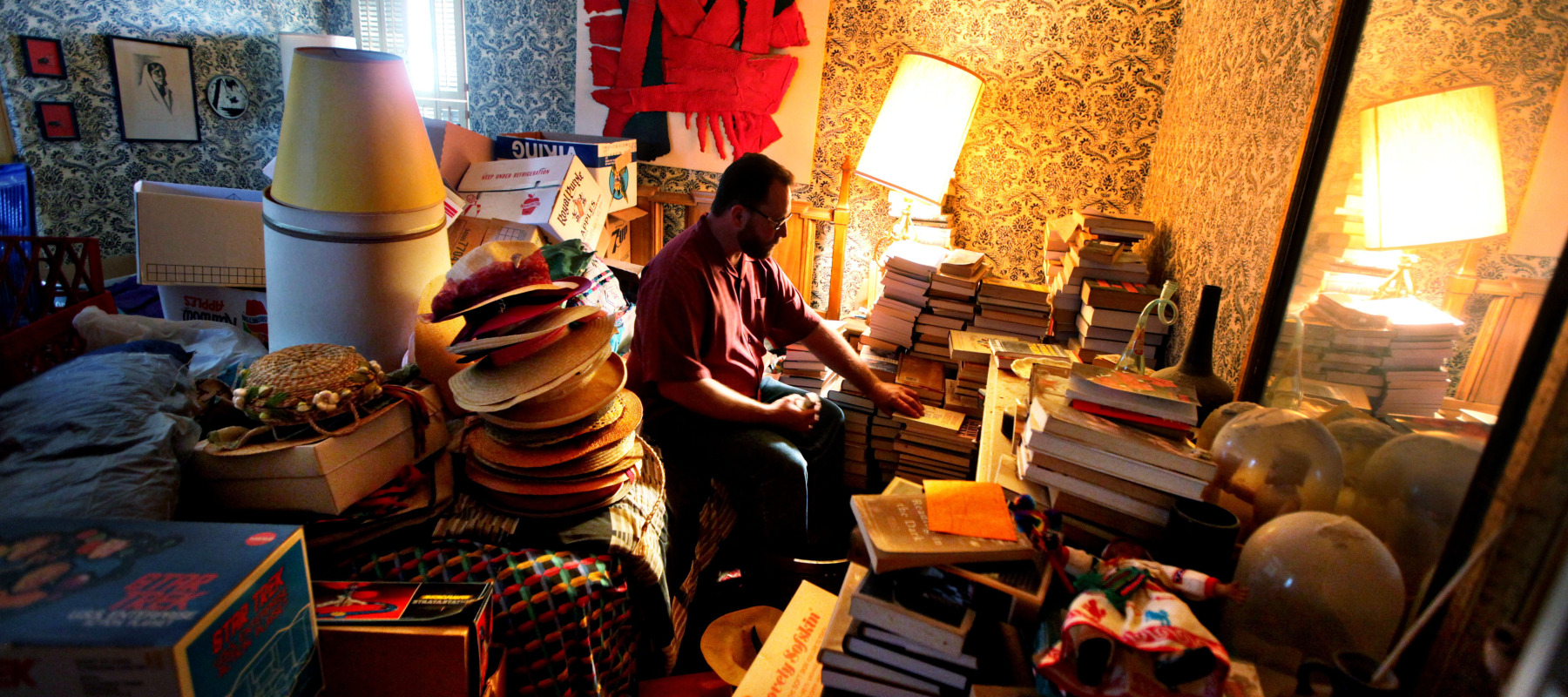 Man sits on bed in room surrounded by stuff, looking down.