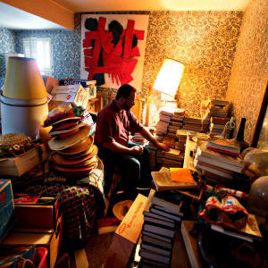 Man sits on bed in room surrounded by stuff, looking down.