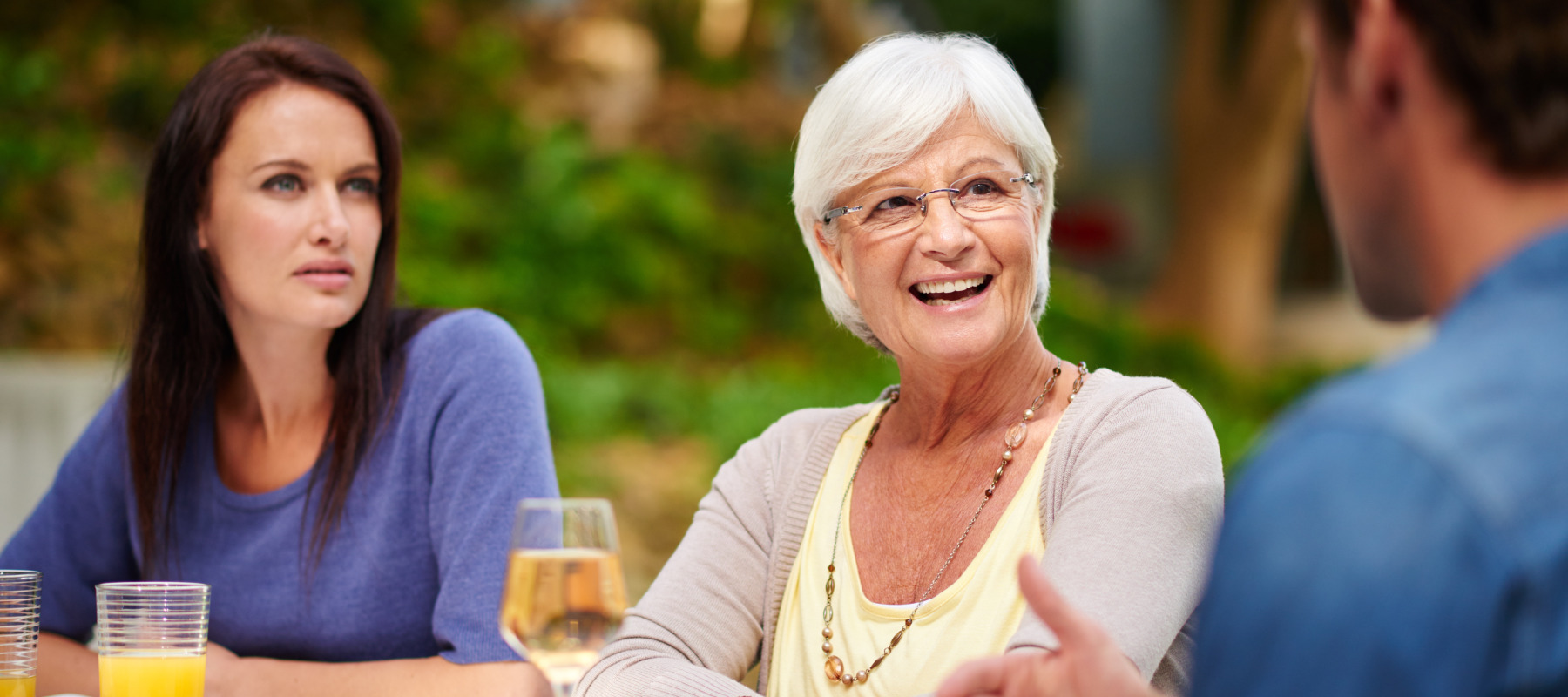 Family sitting around outdoor dining table. Older woman centred and looking towards a man, smiling. Frowning woman behind her.