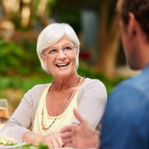 Family sitting around outdoor dining table. Older woman centred and looking towards a man, smiling. Frowning woman behind her.