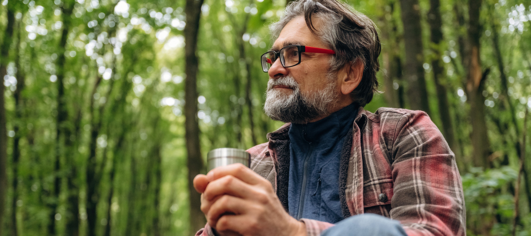 Man looks off into the distance while sitting in the woods, holding a cup.