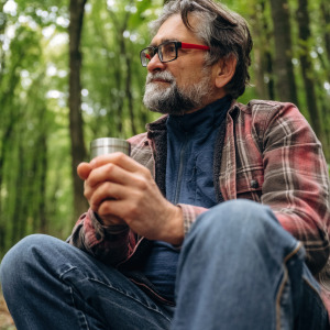 Man looks off into the distance while sitting in the woods, holding a cup.