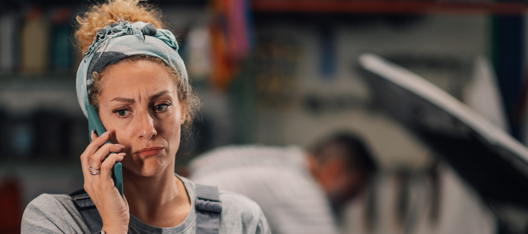 Woman on the phone at a mechanic shop, looking upset, while man works on car in the background.
