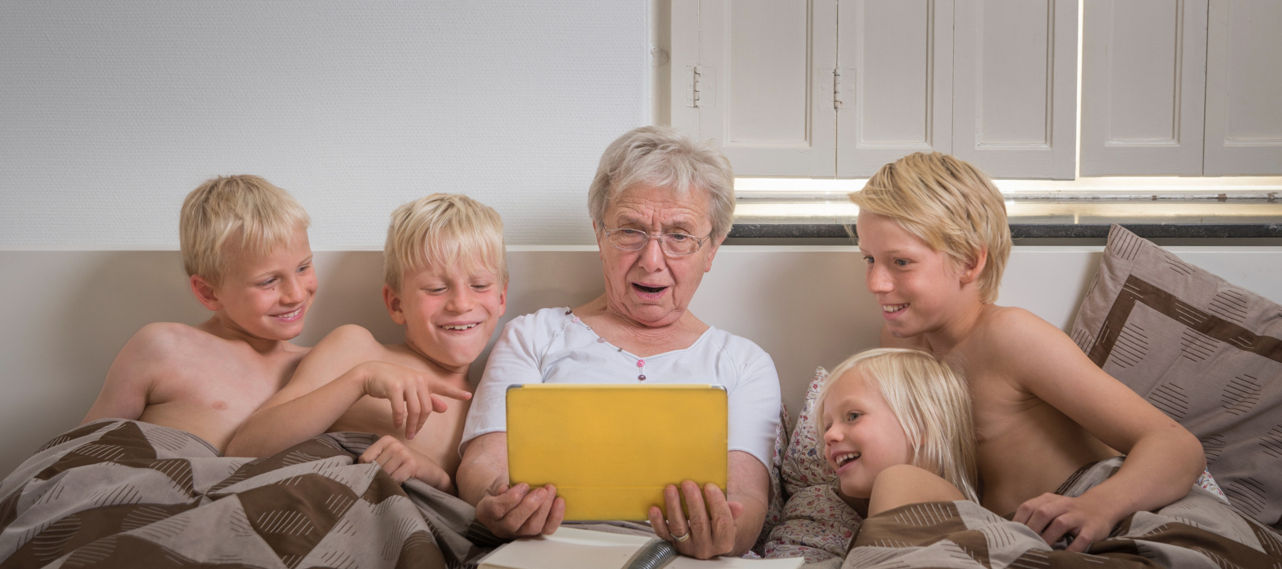 Grandmother sits in bed, looking shocked, surrounded by grandkids laughing.