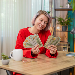 Young white woman counts hundred dollar bills at her kitchen table.