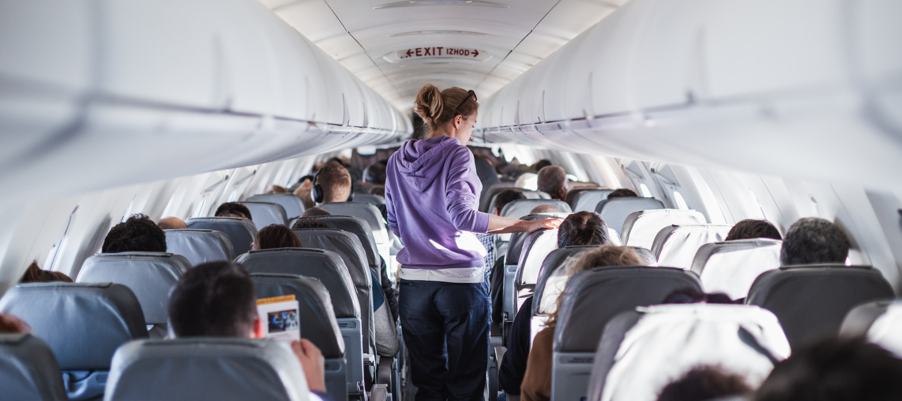 Interior of airplane with passengers in their seats and a female traveler walking the aisle.