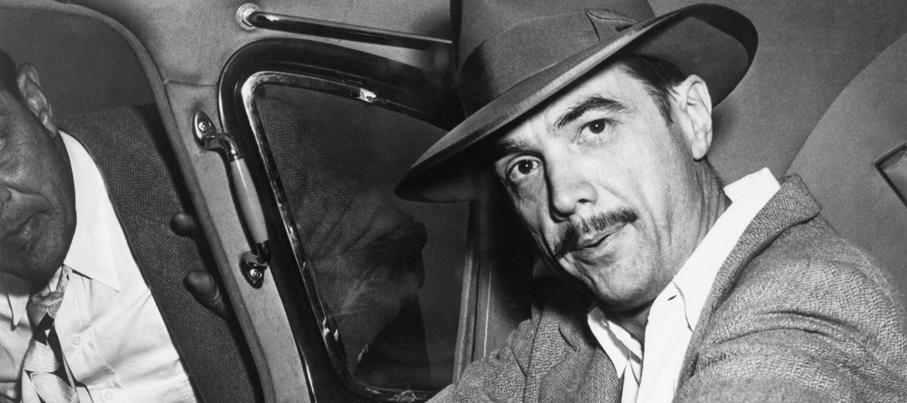 Howard Hughes looking directly at the camera, sitting in the backseat of a car.