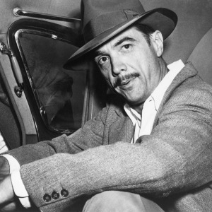 Howard Hughes looking directly at the camera, sitting in the backseat of a car.