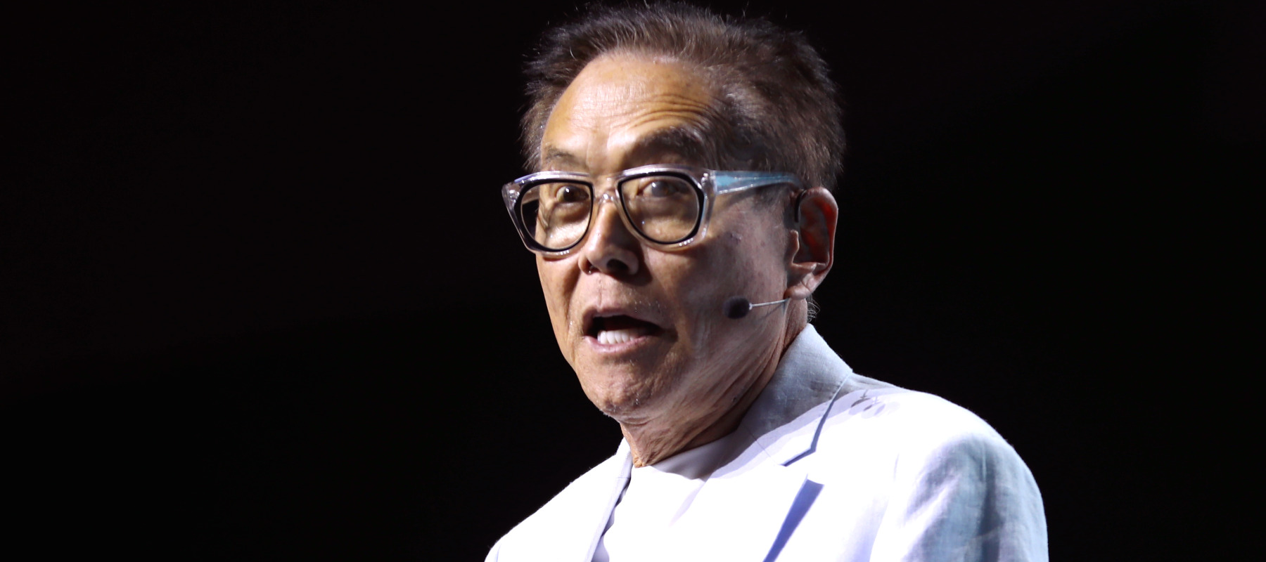 Robert Kiyosaki stands on a dark stage in a bright white suit jacket.
