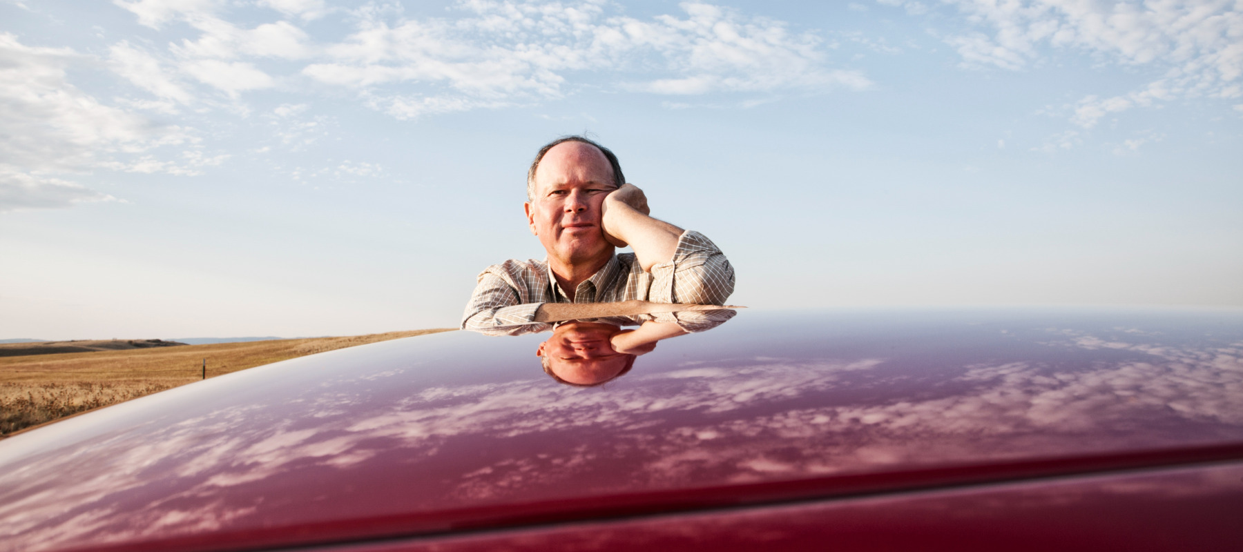 Man seen leaning on the top of a red car, looking directly at the camera.