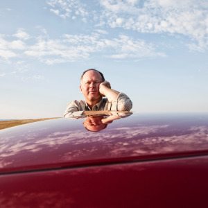 Man seen leaning on the top of a red car, looking directly at the camera.