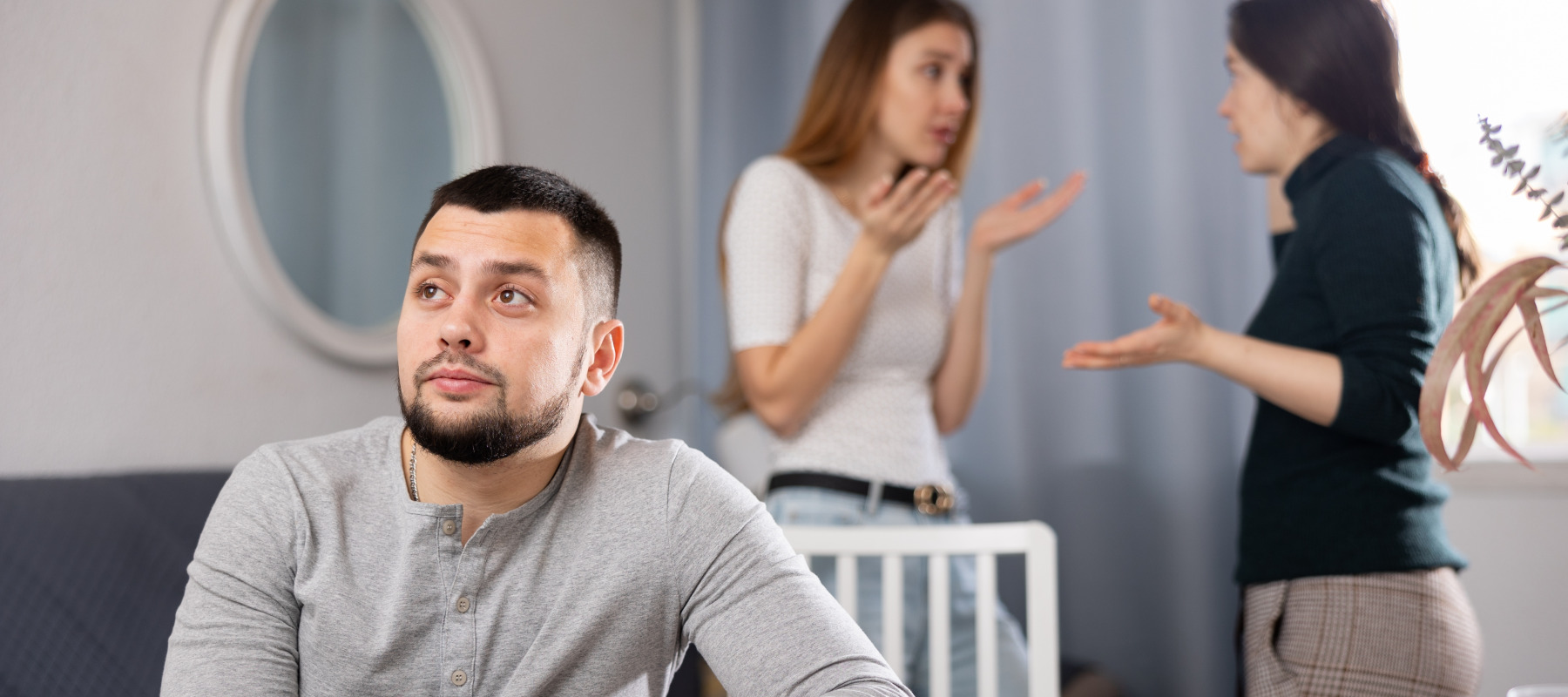 A displeased man sits facing away two female relatives who are fighting.