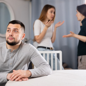 A displeased man sits facing away two female relatives who are fighting.