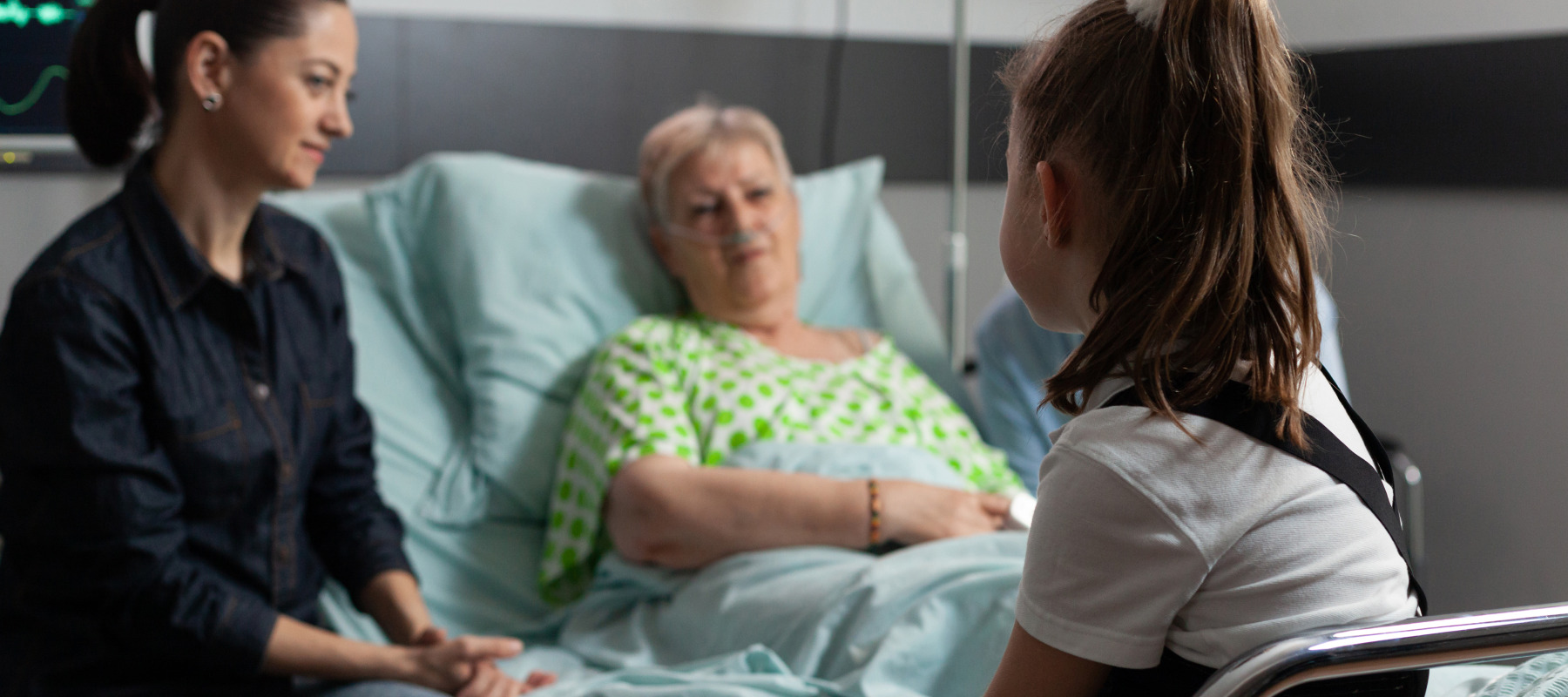 An elderly woman lies in a hospital bed surrounded by her family, with her granddaughter in the foreground.