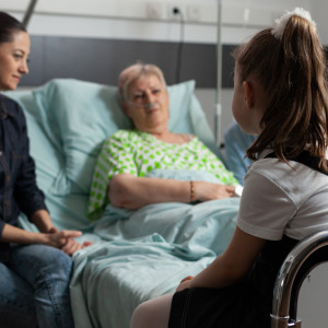 An elderly woman lies in a hospital bed surrounded by her family, with her granddaughter in the foreground.