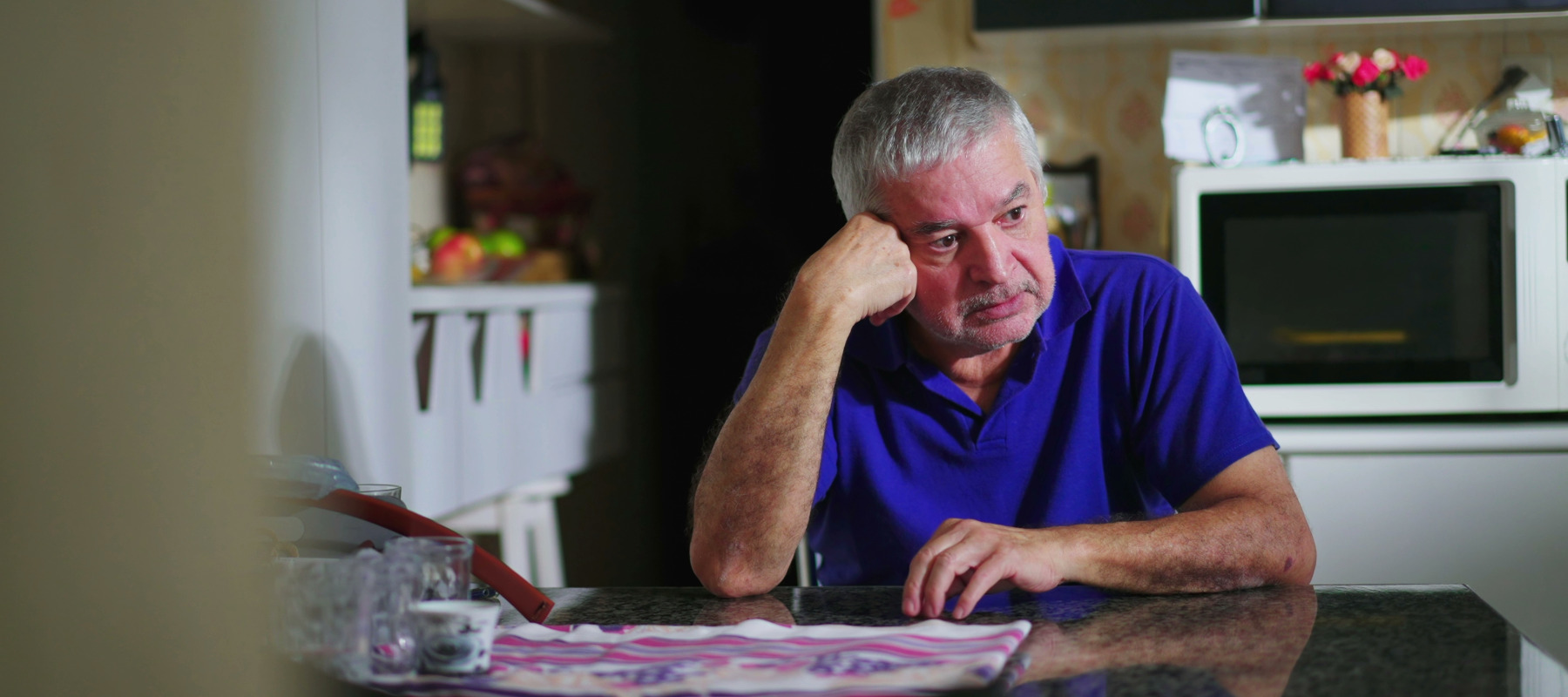A middle-aged man sits at his kitchen table looking pensive.
