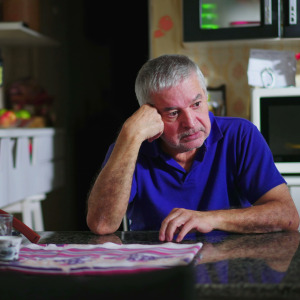A middle-aged man sits at his kitchen table looking pensive.