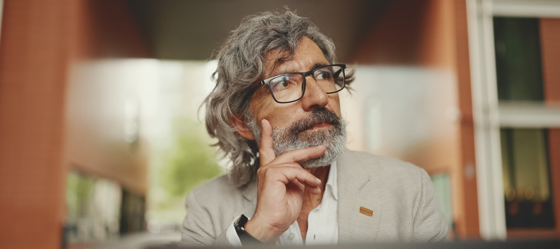 An adult man with gray hair and glasses sits outdoors in what appears to be a city setting, looking off to the side.