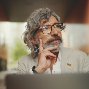 An adult man with gray hair and glasses sits outdoors in what appears to be a city setting, looking off to the side.
