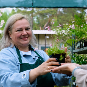 A middle-aged woman hands a plant to a customer at a garden center.