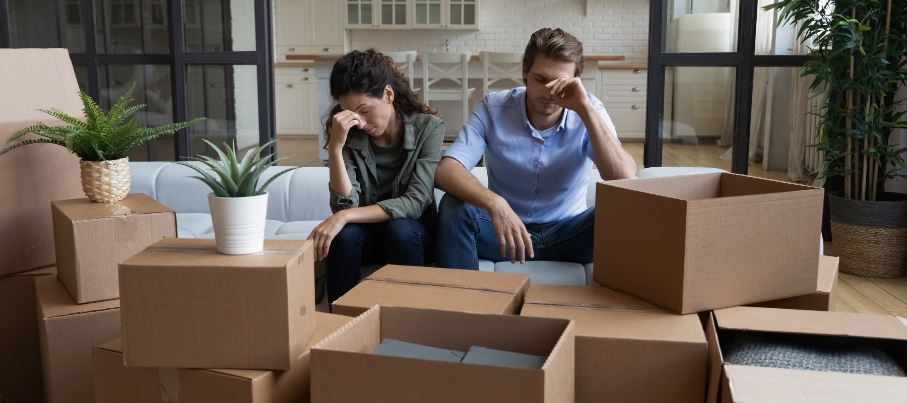 A frustrated couple sits on a couch with moving boxes around them.