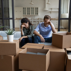A frustrated couple sits on a couch with moving boxes around them.