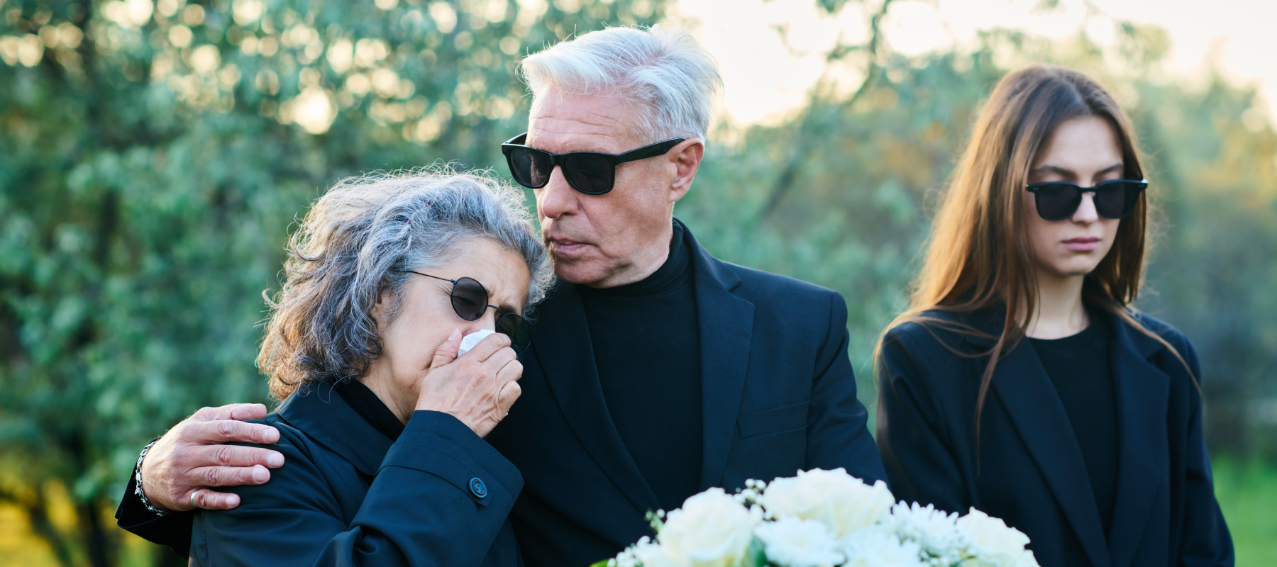 Older man seen at the side of a grave, with his arm around a woman crying and another girl by his side.
