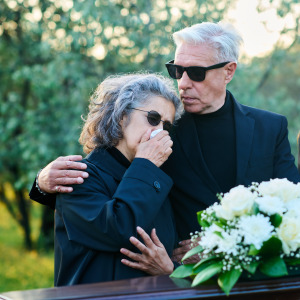 Older man seen at the side of a grave, with his arm around a woman crying and another girl by his side.