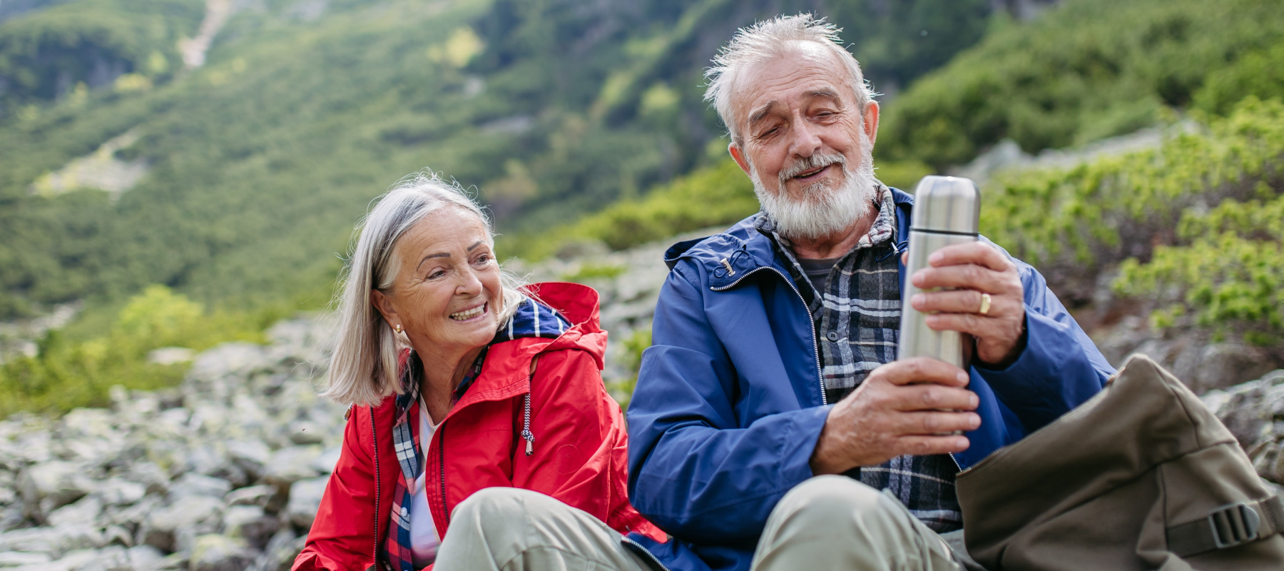 An older couple on a hikes take a break with a thermos as mountains loom behind them.