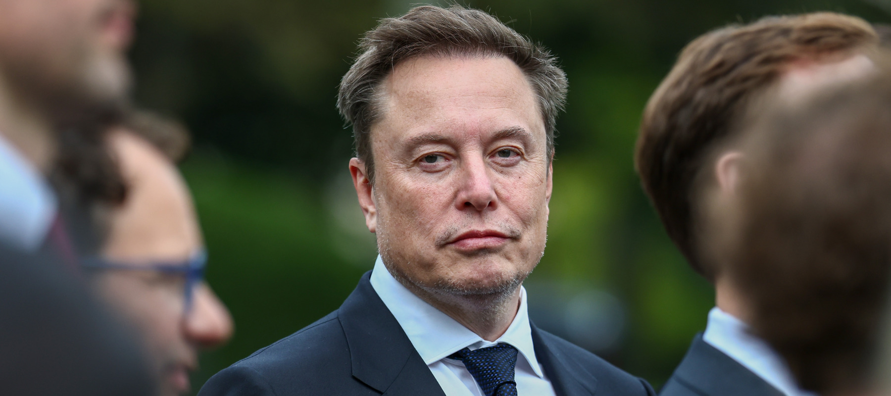 Elon Musk, wearing a suit and tie, looks directly at the viewer through a busy crowd.