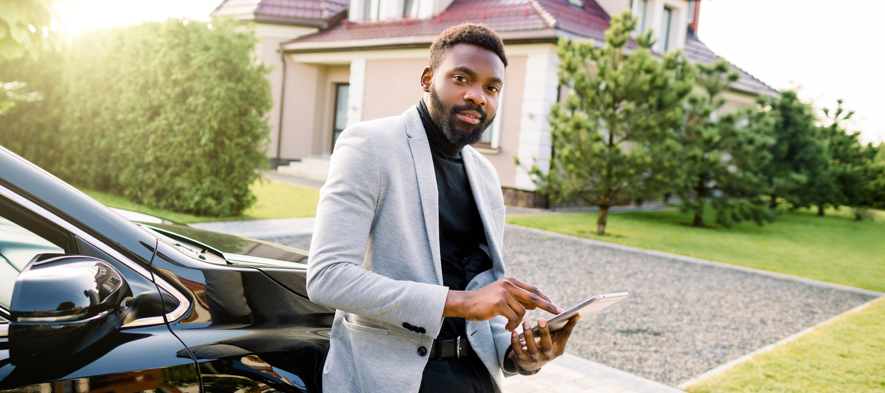 A sharply-dressed man with a neatly trimmed beard stands leaning against a black vehicle, using a tablet.