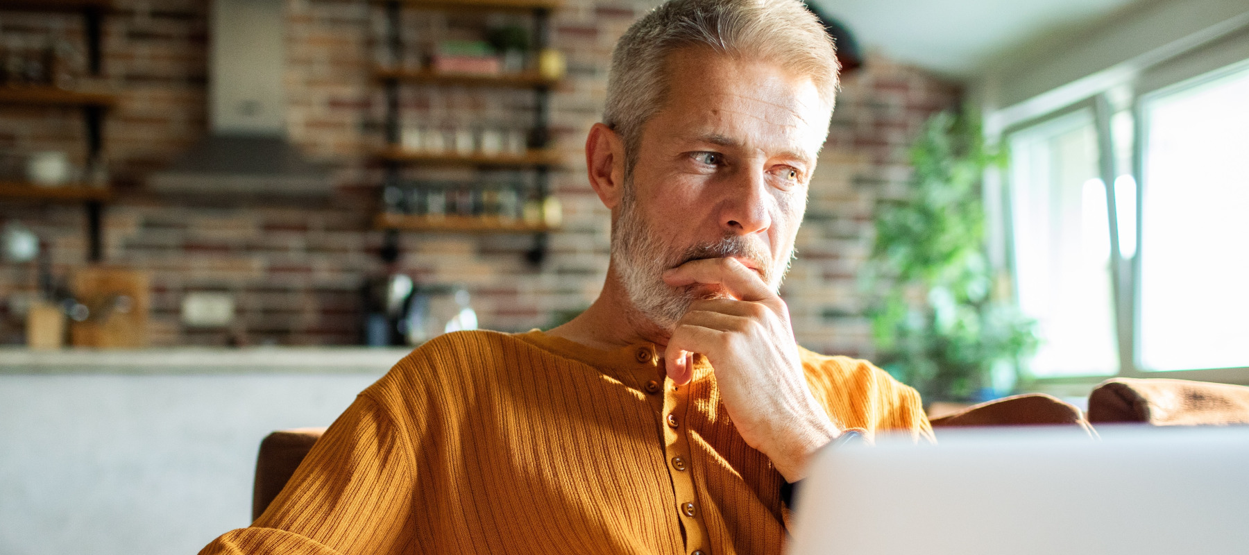 Middle-aged man thinking while using laptop at home.