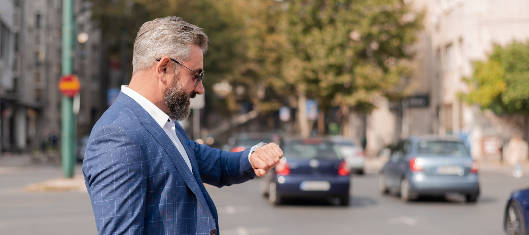 A businessman in a dapper suit looks at his watch while crossing the road.