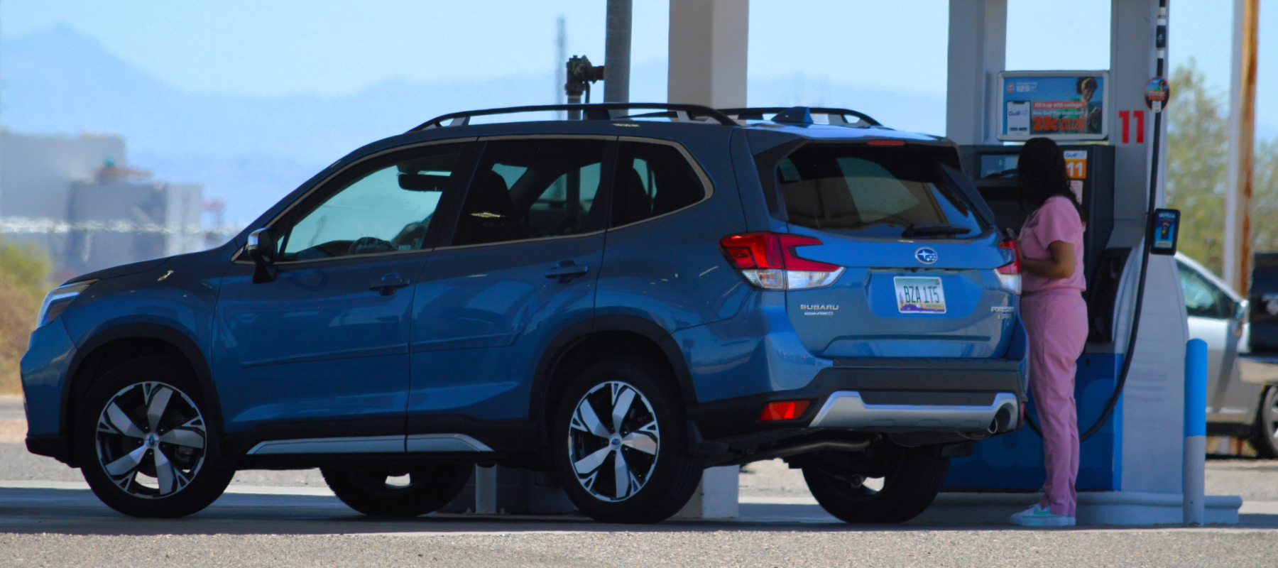 Woman in pink nurse scrubs pumps gas into her Subaru SUV.