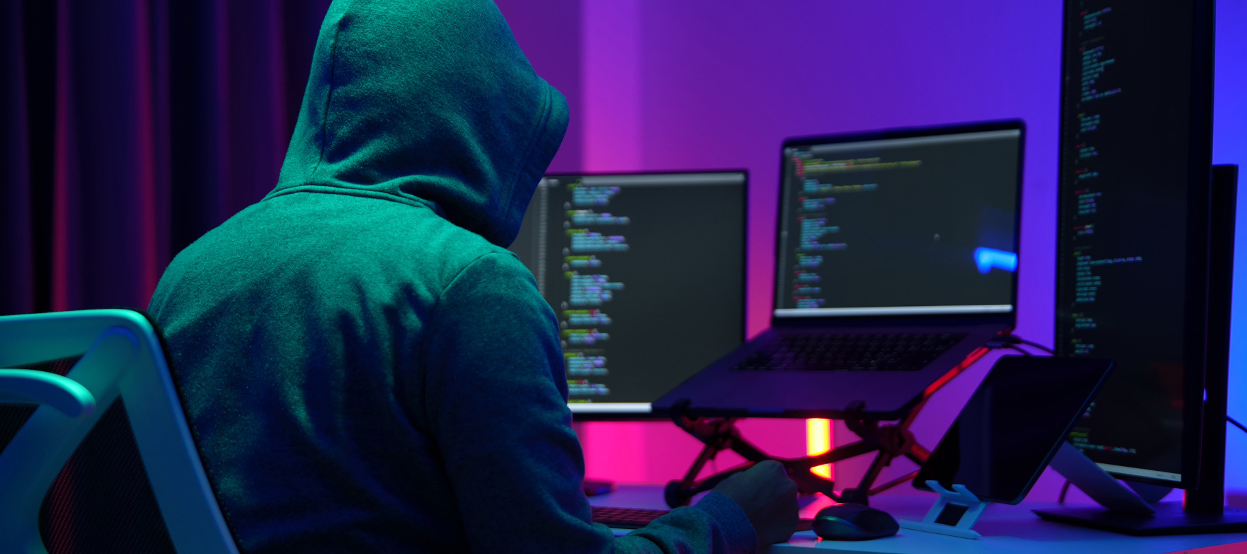 A hood man sits at a computer terminal with a mobile phone in his hand.