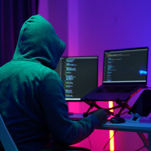 A hood man sits at a computer terminal with a mobile phone in his hand.