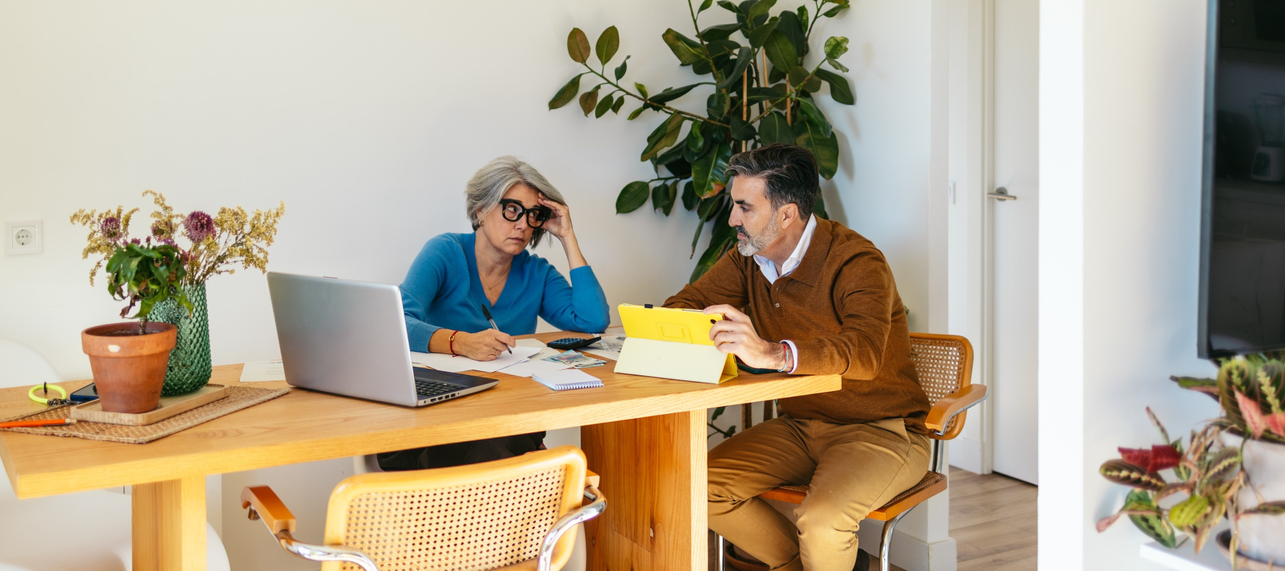 Middle aged couple sit at dining room table together, shooting concerned looks at each other while going over paperwork.