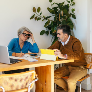 Middle aged couple sit at dining room table together, shooting concerned looks at each other while going over paperwork.