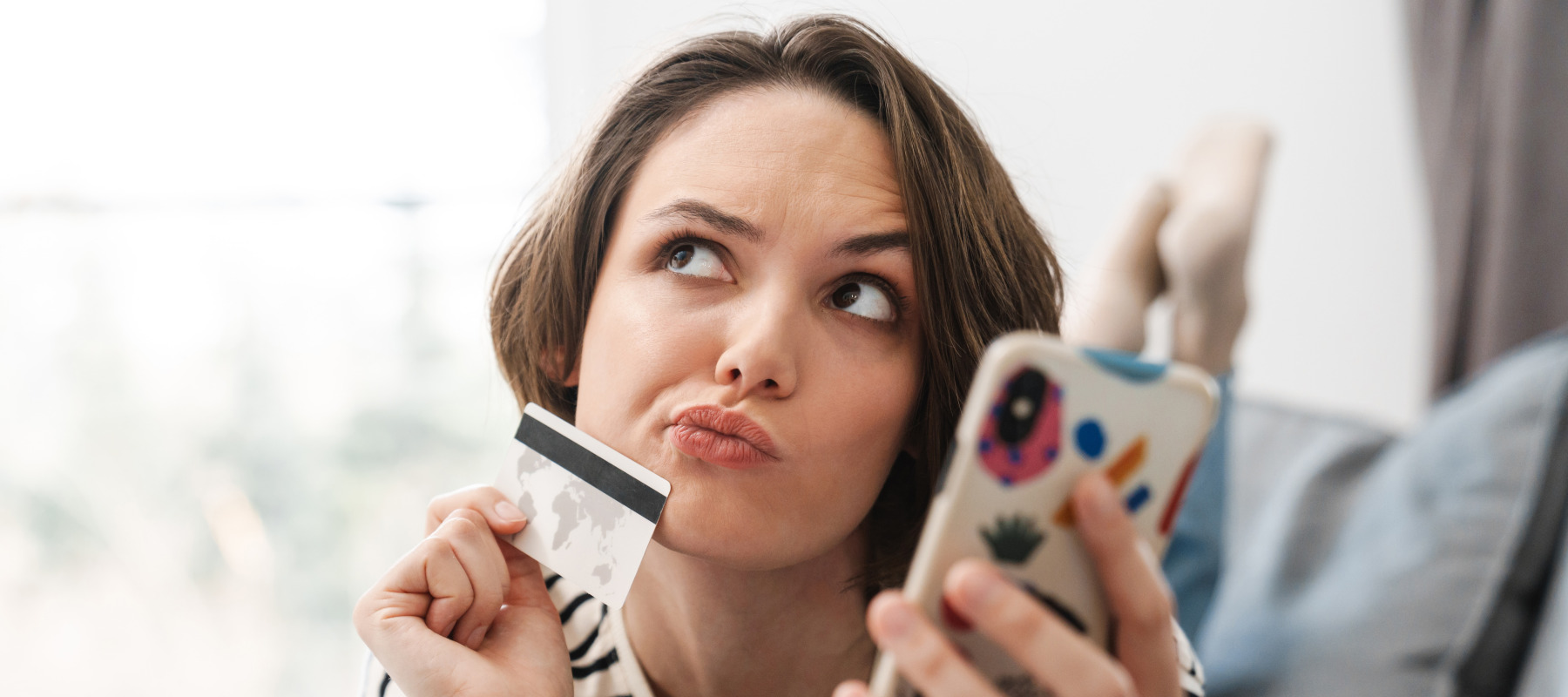 A young woman with short wavy brown hair lays on a bed looking upwards and holding a credit card and a phone.
