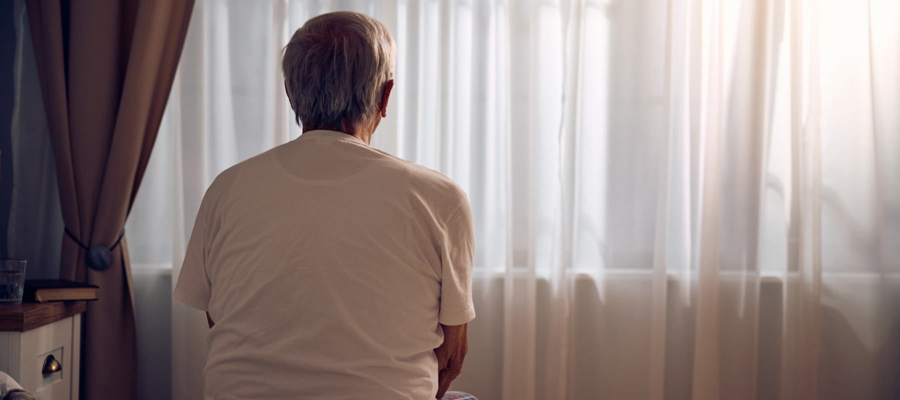Elderly white man dressed in loungewear looks outside of his bedroom window.