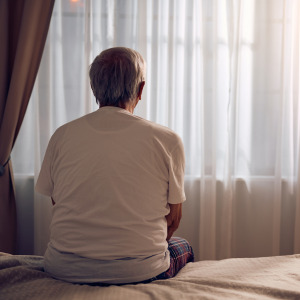 Elderly white man dressed in loungewear looks outside of his bedroom window.