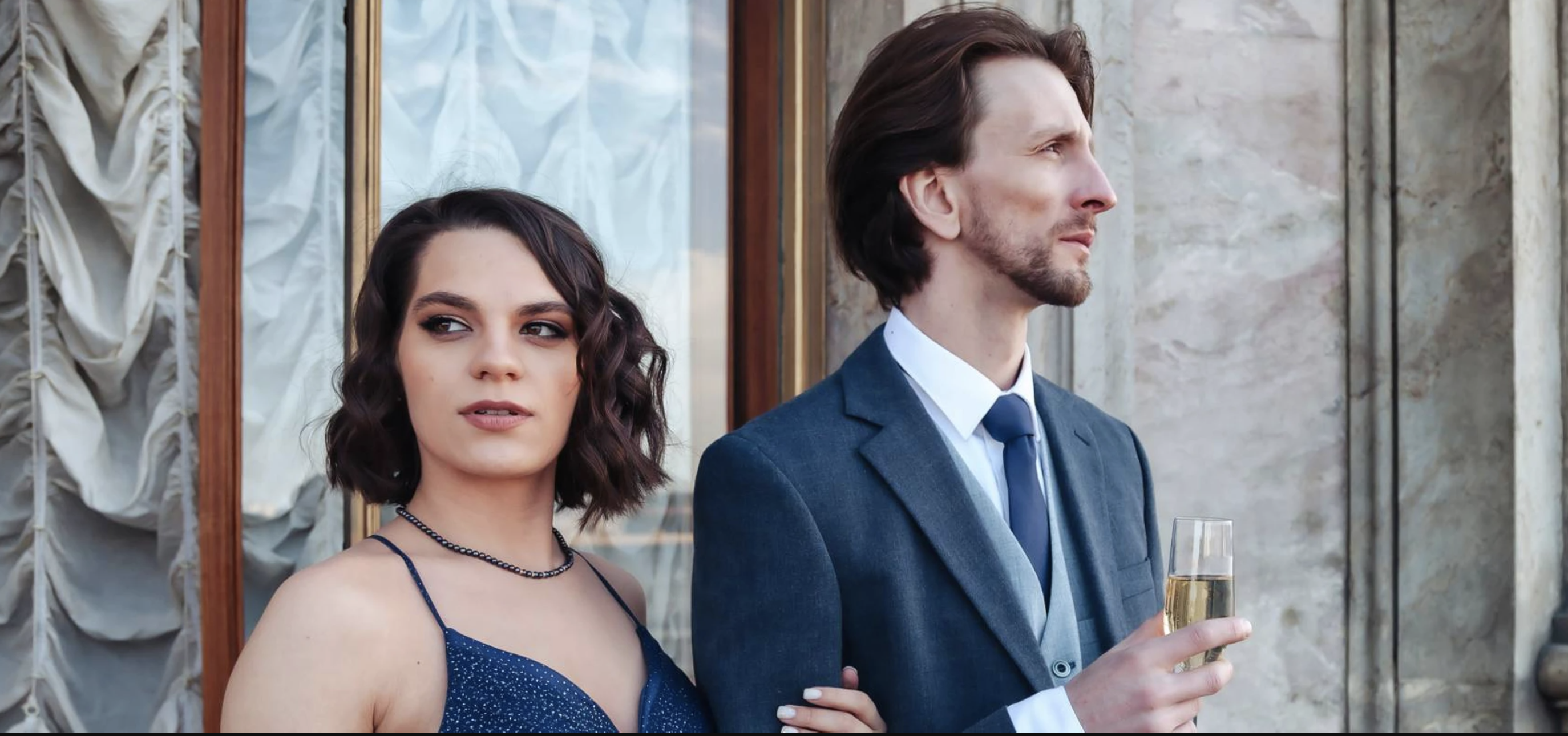 A wealthy young couple pose outside a mansion.