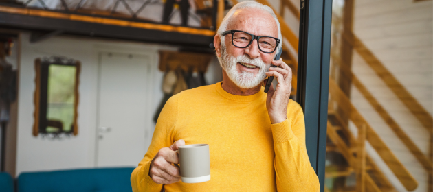 Man in his 50s-60s standing with a cup of coffee in his hand and a phone to his ear.
