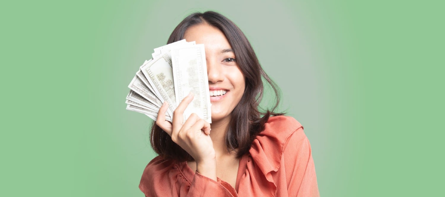 Smiling woman holding up cash covering her face with a green background.